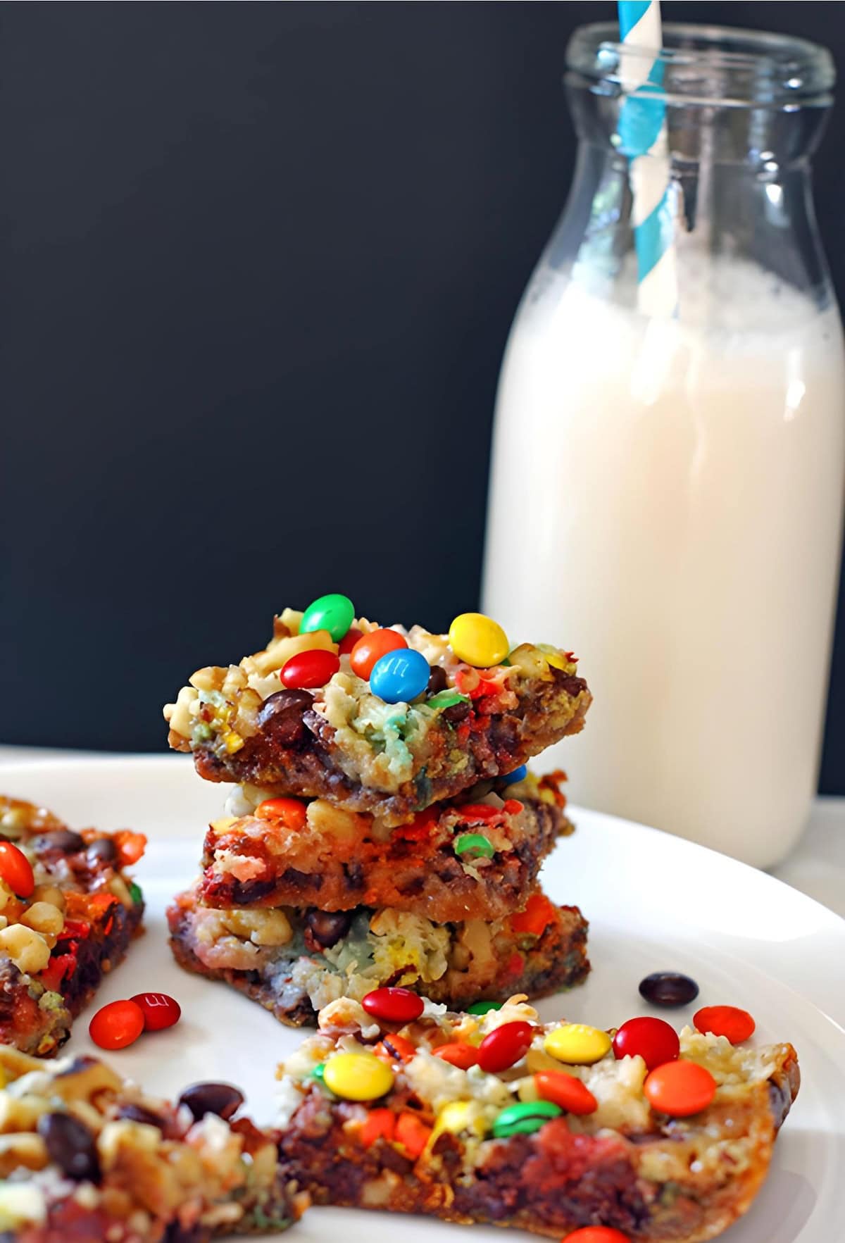 Four 7-layer cookie bars with nuts, coconut, chocolate chips, and mini m and ms stacked on a white plate with a glass of milk.