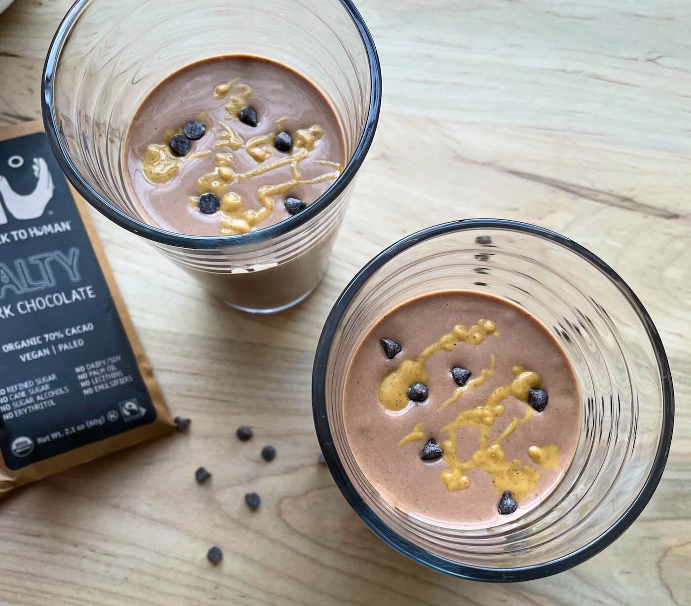 Two chocolate peanut butter banana smoothies with peanut butter drizzled over the top and chocolate chips sprinkled around.