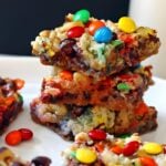 Gooey magic cookie bars with nuts, coconut, chocolate chips, and mini m and ms stacked on a white plate with a glass of milk.
