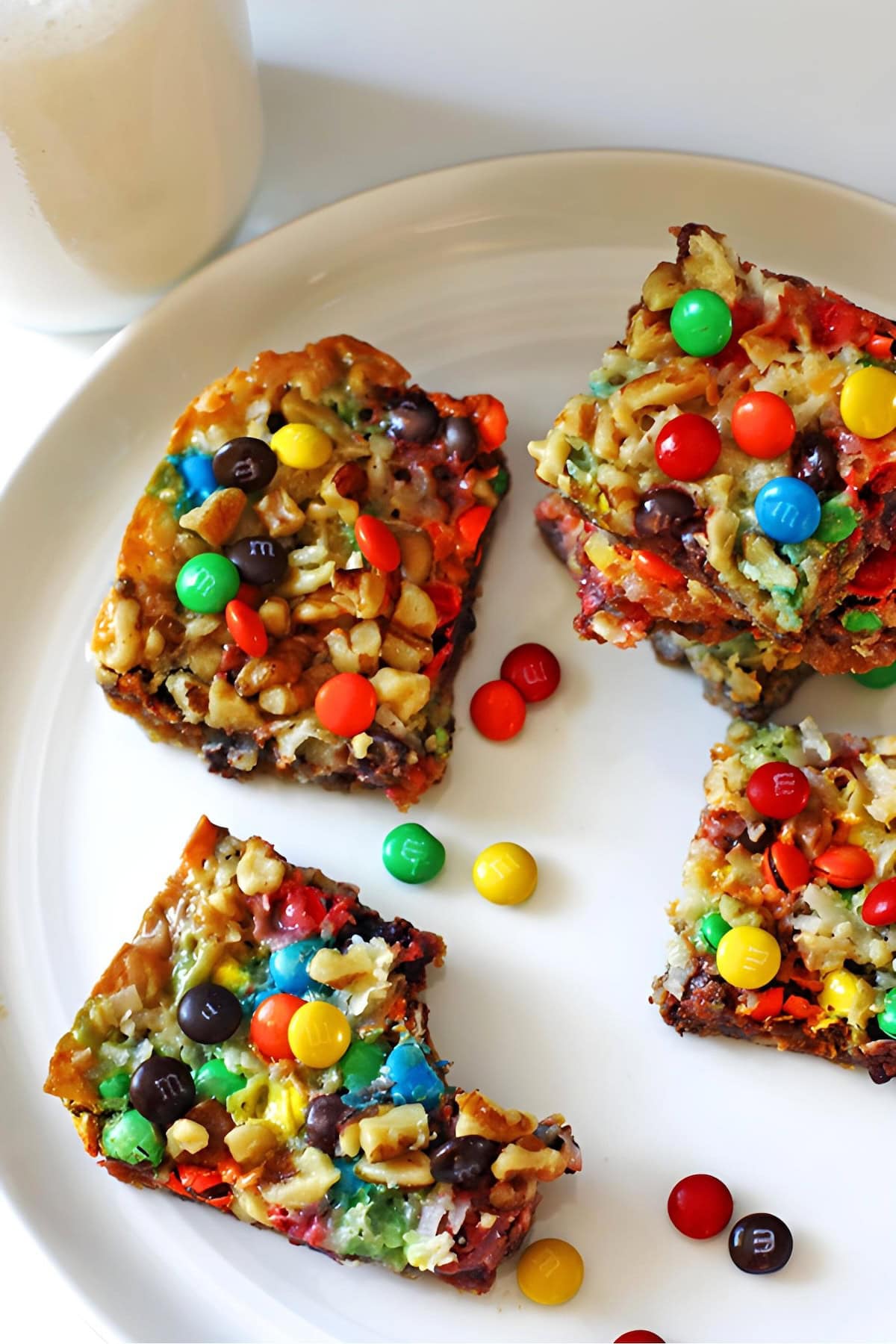 Four magic cookie bars with walnuts, coconut, chocolate, and mini m and ms on a white plate with a glass of milk.