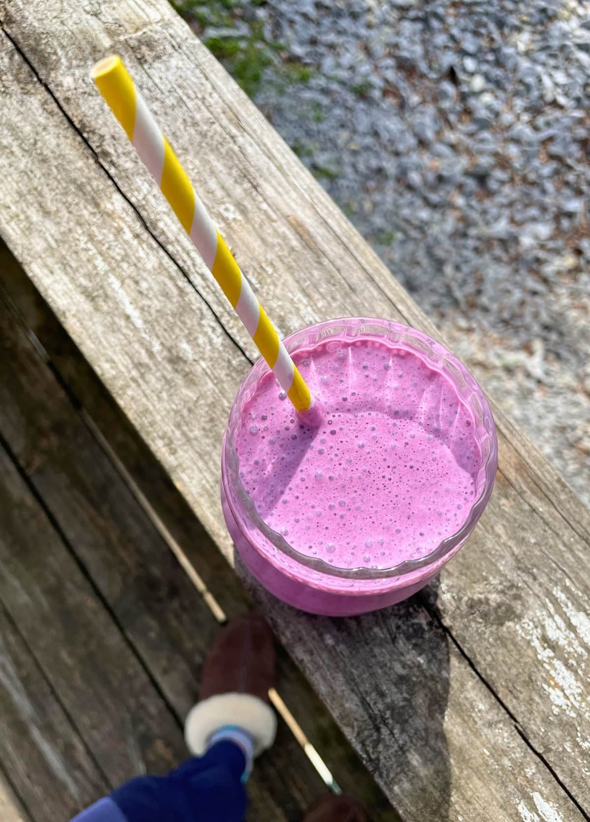 A blueberry pineapple smoothie with a white and yellow striped straw on a railing overlooking a gravel driveway.