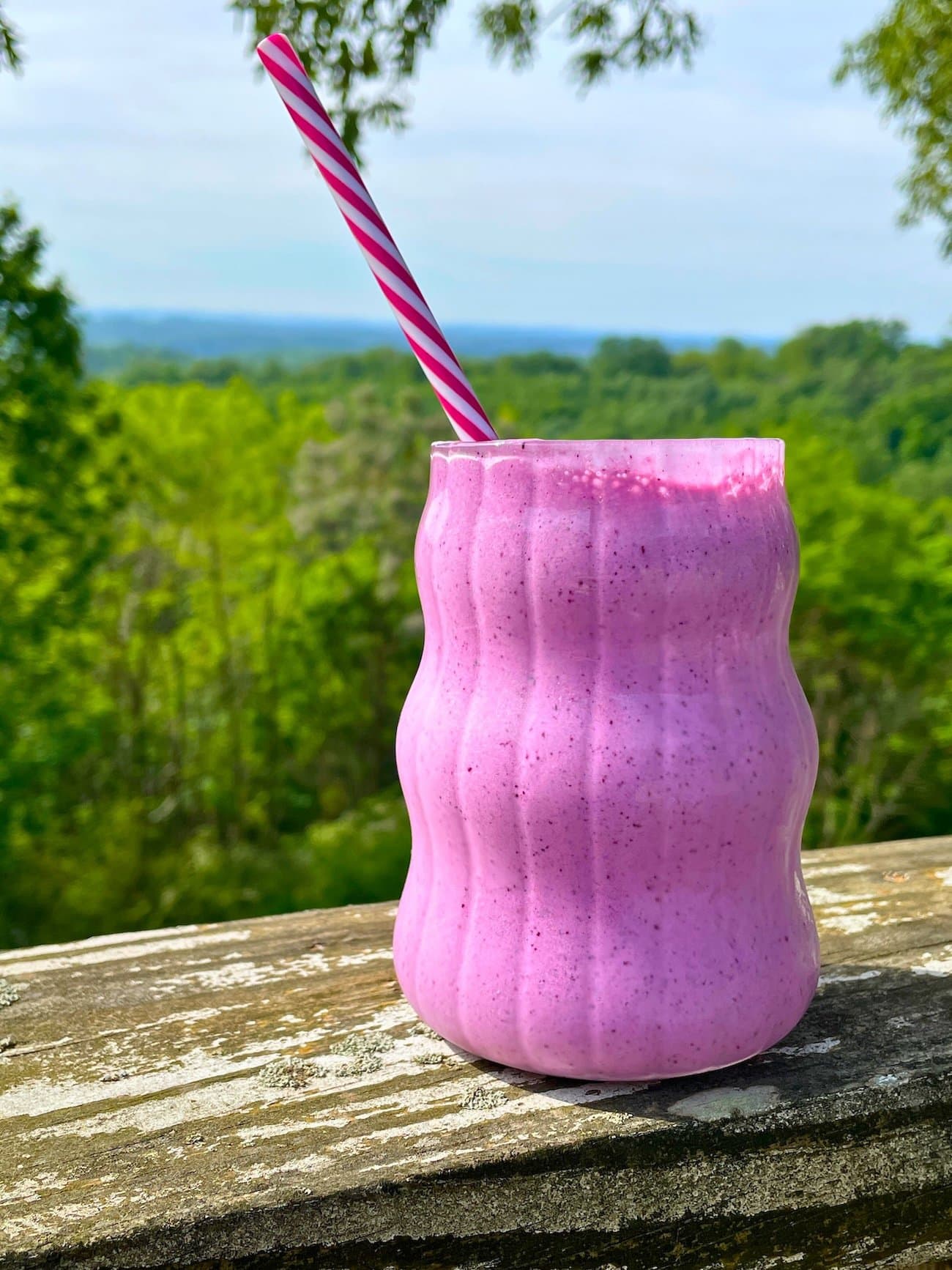 A creamy blueberry pineapple smoothie in a glass with a pink straw on a wood railing outside with trees behind it.