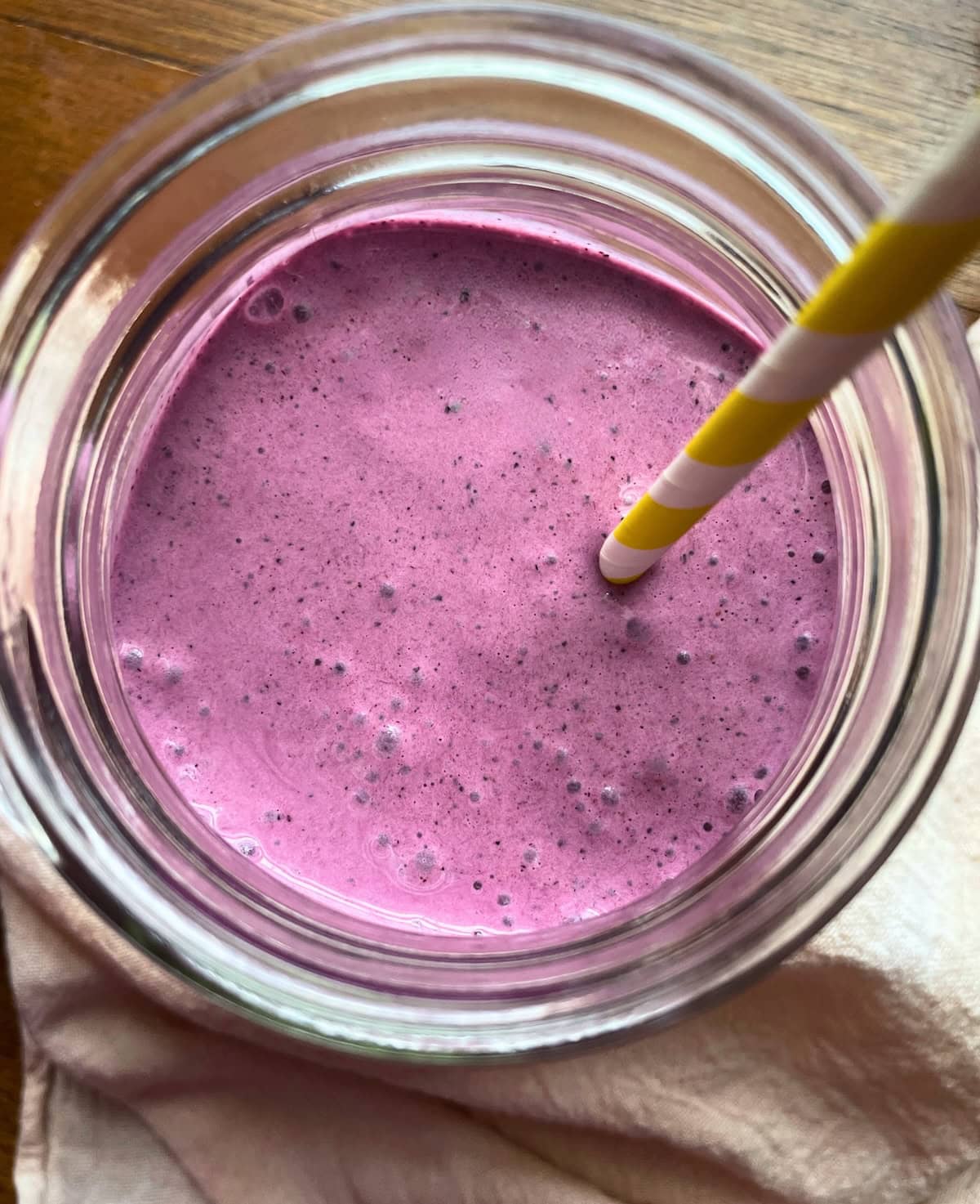 A creamy blueberry smoothie with pineapple, banana, yogurt, and spinach in a mason jar with a yellow and white striped straw viewed from the top.