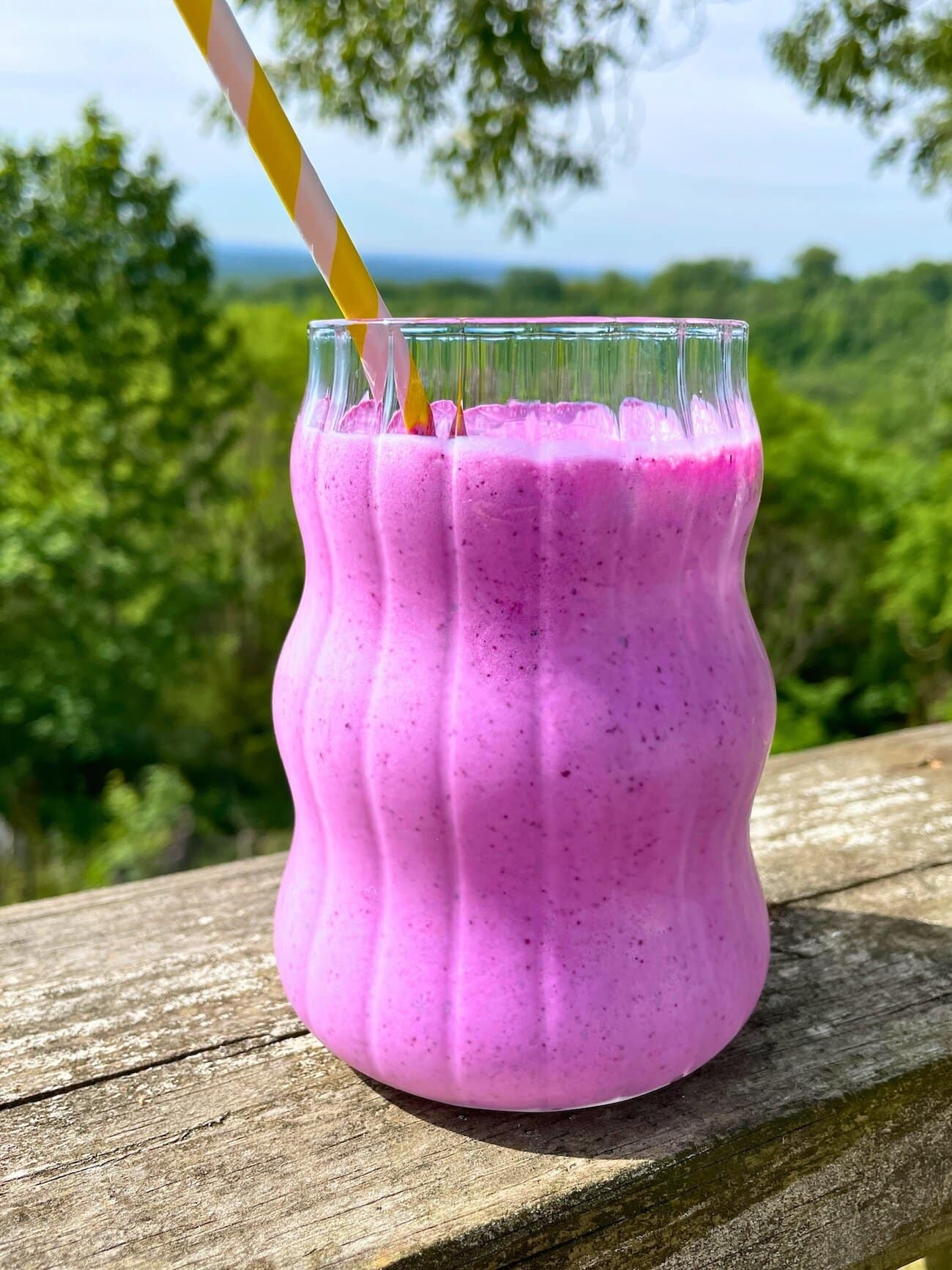 Blueberry pineapple smoothie in a glass with a yellow straw on a wood railing outside with trees behind it.