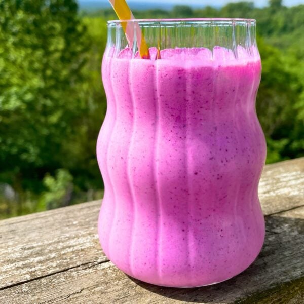 Blueberry pineapple smoothie in a glass with a yellow straw on a wood railing outside with trees behind it.