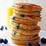 A stack of fluffy blueberry pancakes with maple syrup dripping down the side and blueberries scattered around.