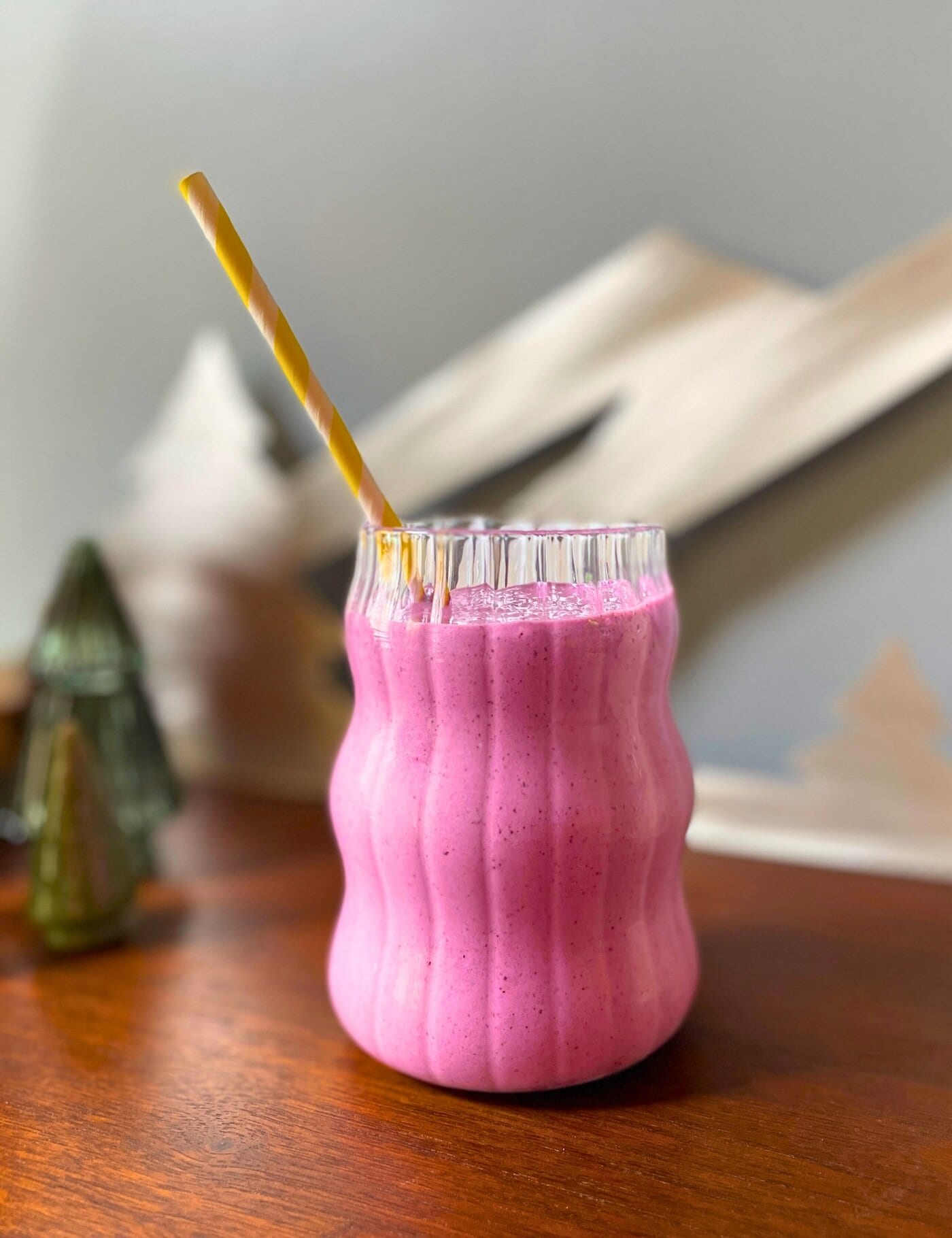 A blueberry pineapple smoothie in a glass with a yellow straw on a wood cabinet with a mountain sign and glass trees behind it.