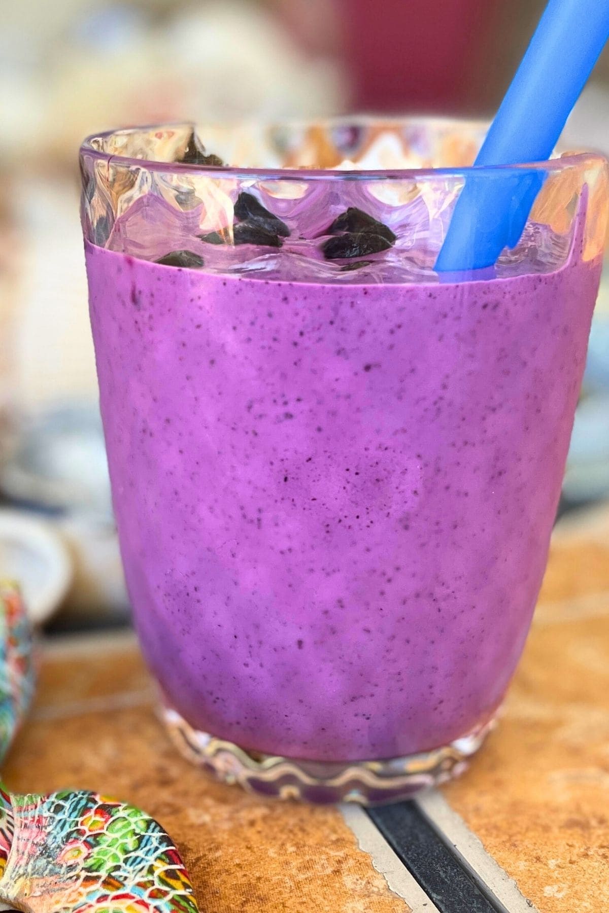 A bluberry banana and peanut butter smoothie in a clear glass on a tiled background with a blue straw and shells behind it.