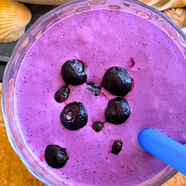 A bluberry banana and peanut butter smoothie with blueberries on top on a tiled background with a blue straw and shells off to the side.