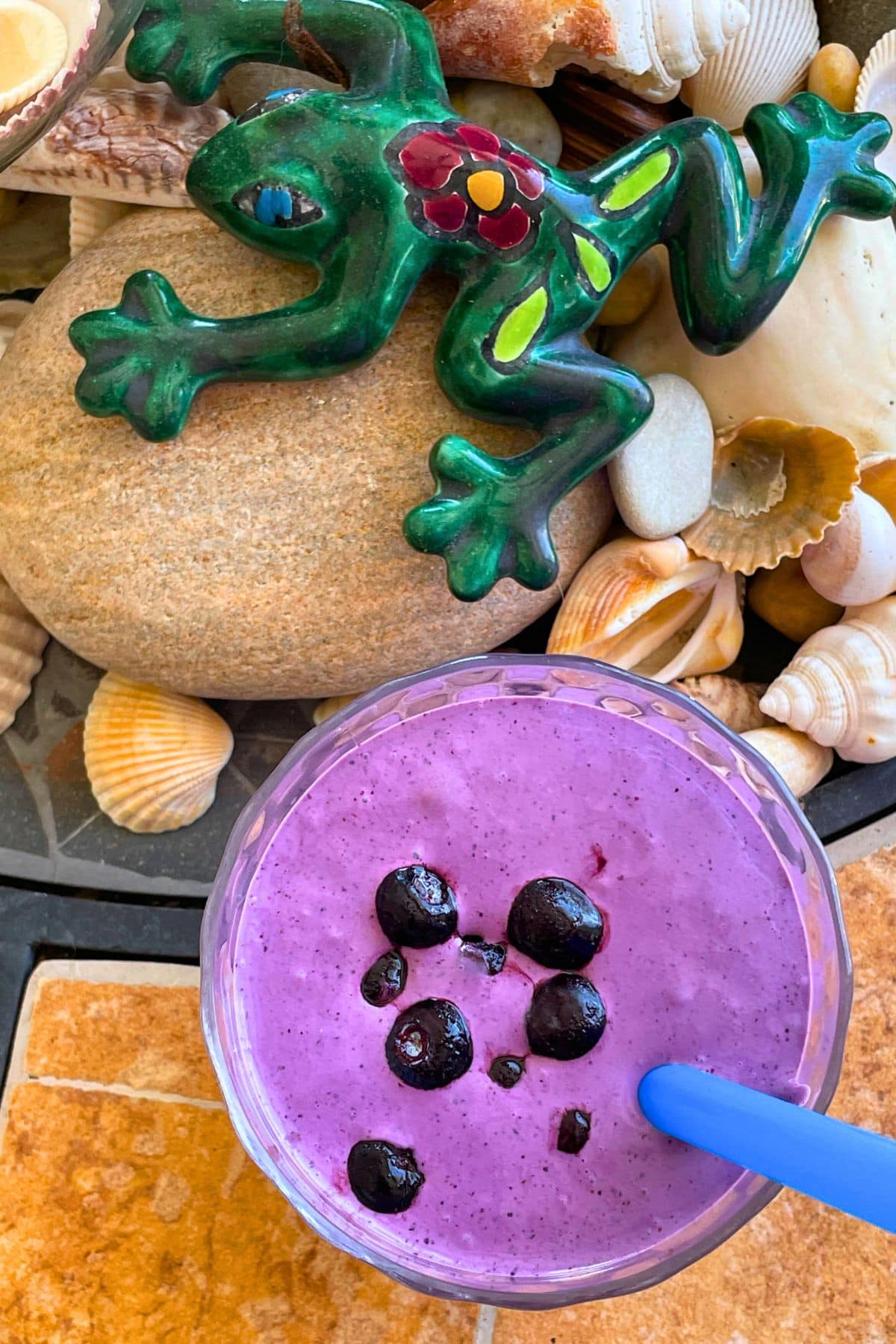 A bluberry banana and peanut butter smoothie with blueberries on top on a tiled background with a blue straw and a patterned frog and shells off to the side.