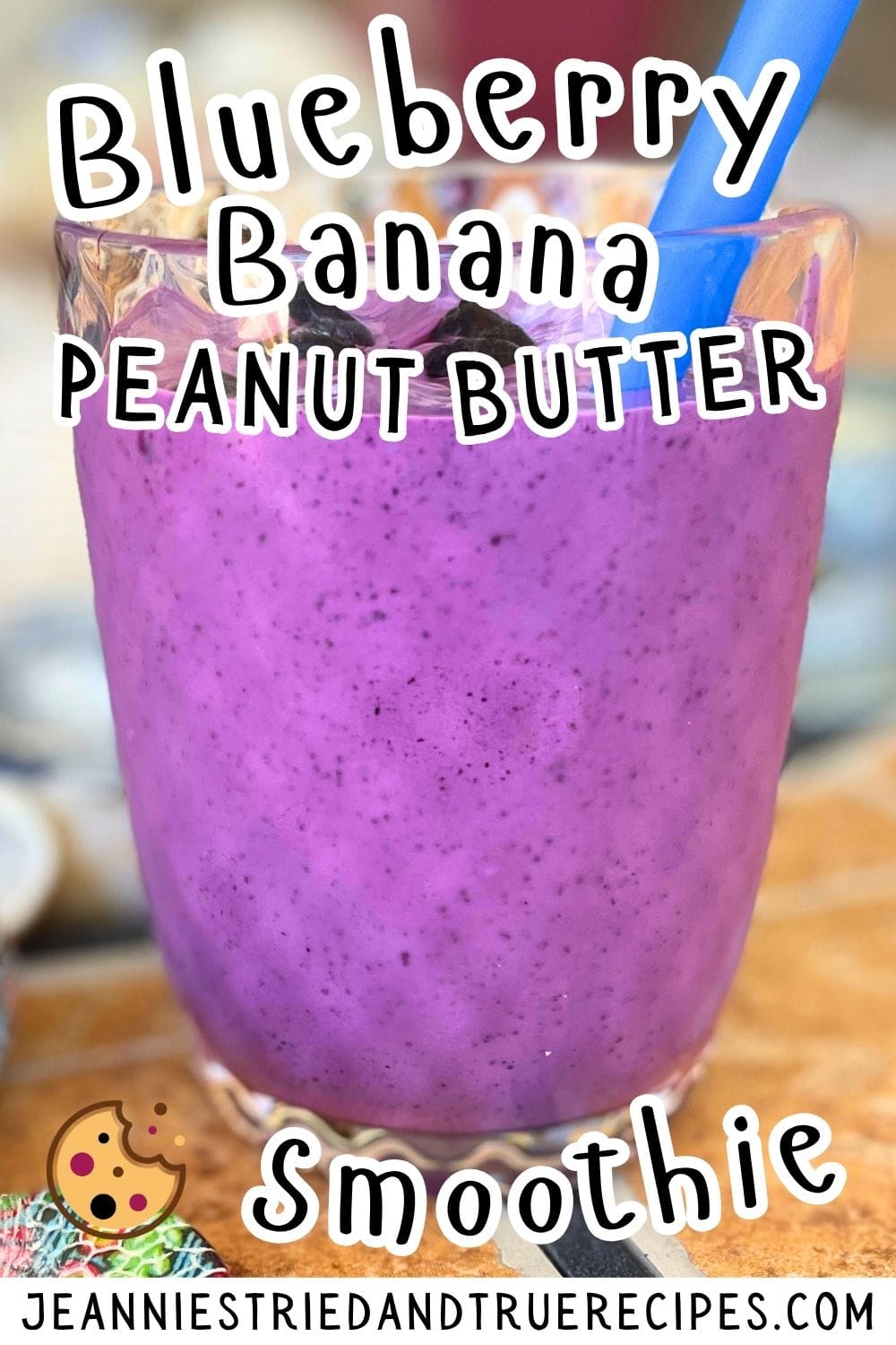 A pin image of a bluberry banana and peanut butter smoothie in a clear glass on a tiled background with a blue straw and shells behind it.