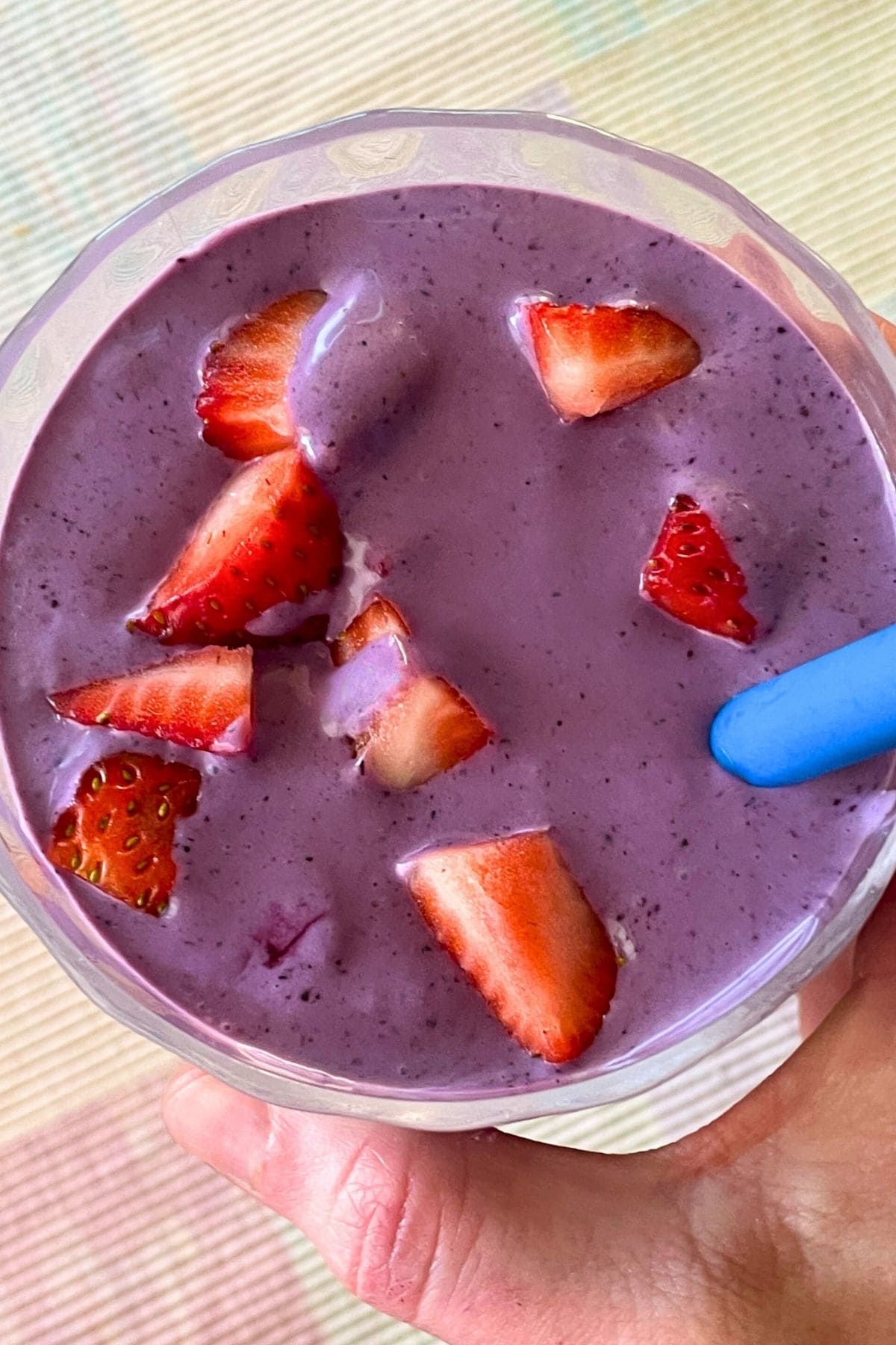 A blueberry banana and peanut butter smoothie with a blue straw held in a hand in a clear galss topped with some diced strawberries.